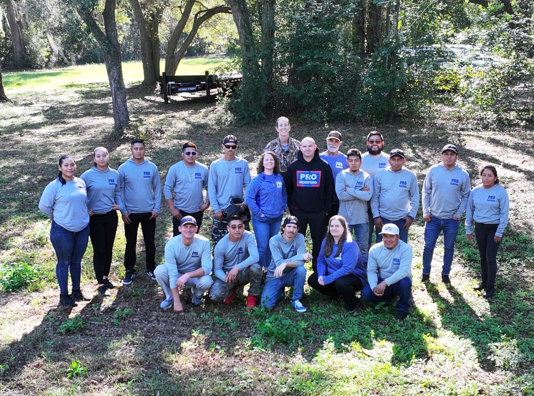 A group of 18 people, most wearing matching gray shirts, pose outdoors on grass with trees and a picnic table in the background.