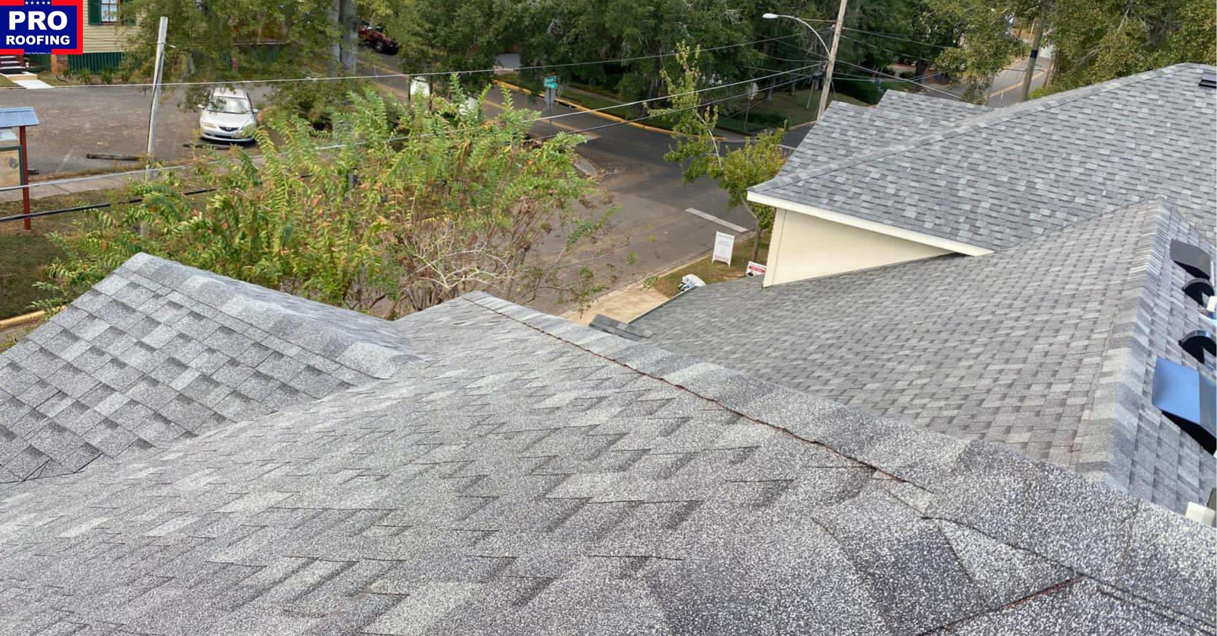 Gray shingle roof viewed from above, with trees, a street, parked cars, and neighboring buildings visible in the background; Pro Roofing sign in upper left corner.